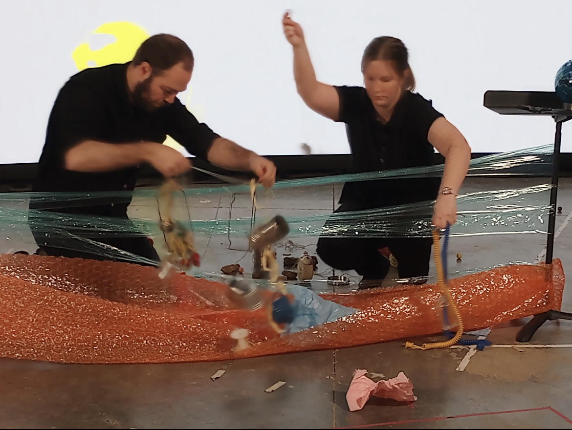 picture of performers interacting with an uncanny collection of objects including stretched plastic wrap